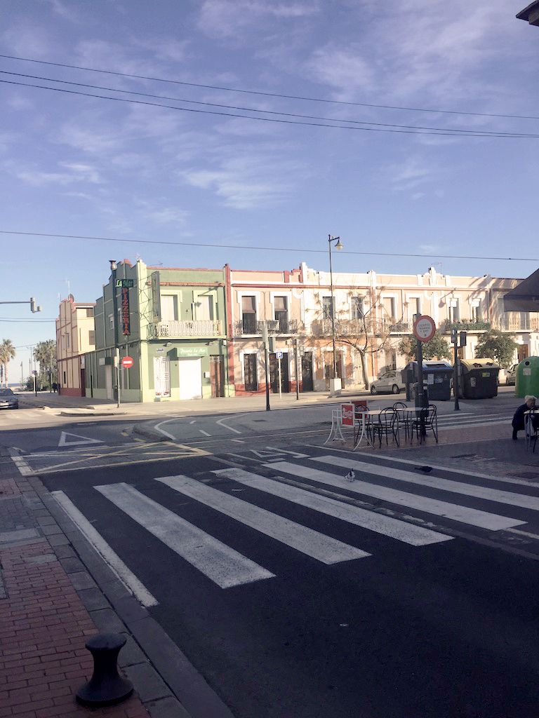 In the foreground of the image is a zebra crossing. Beyond this, a row of beautiful ornate houses, painted in green, orange and peach sit to the right of the image. There is a small table and four chairs to the right of the zebra crossing. A person crouches down near these, almost out of shot. Other than this person there is no one in the image. It appear eerily quiet.