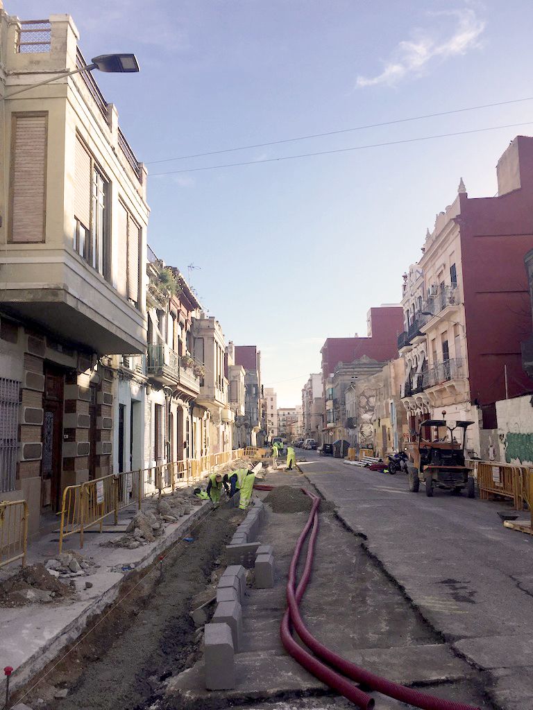 A row of houses sit on either side of this image. The buildings look to be quite grand and beautiful. Running through the middle of the image towards the horizon is the street where reconstrucitve works are being done. The road has been dug up to allow for these repairs, and workmen in high–vis clothing are to the left of the image, undertaking various tasks.
