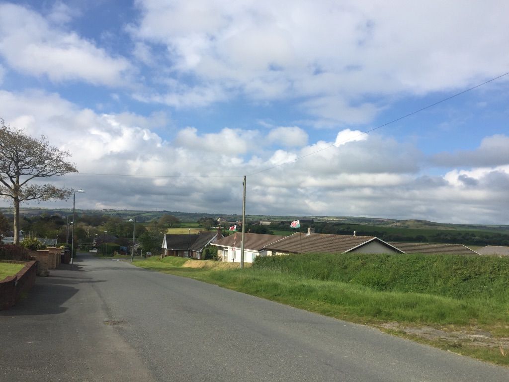 A road runs down the center of the image. To the left are a few bungalows, their roofs just visible beyond a grassy verge. In front of the bungalows are two Welsh flags blowing in the wind.
