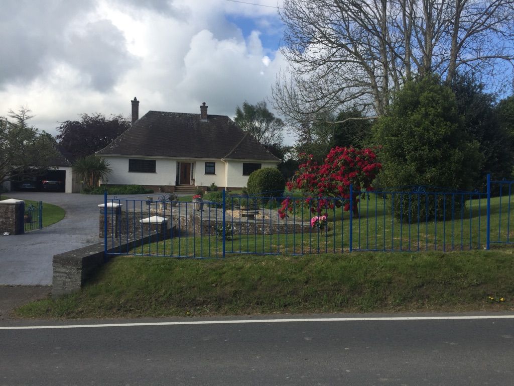 A white bungalow with a greyish–brown roof is in the background of the image. A blue fence enters the frame from the right hand side, spanning three quarters of the image before stopping next to a small brick wall that leads up the driveway to the property. Behind the fence is the front garden. There is a beautiful red flowering bush in bloom next to a rounded green hedge.