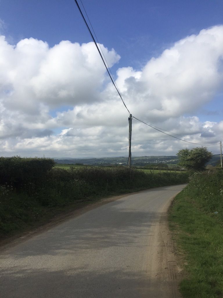 The image is focused on the road in the center. It is between two grassy verges and bends to the right and out of frame. On the left in the distance there is a telegraph pole. The sky is bright blue behind thick white clouds.