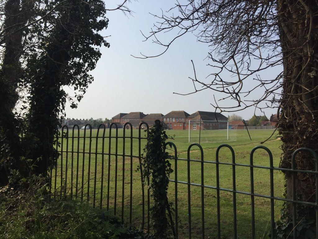 This image shows a group of six red brick buildings, looked at over the top of some metal railings through a gap in the trees. These buildings are just beyond a large green school field. There is a white football goal in view in the right half of the image