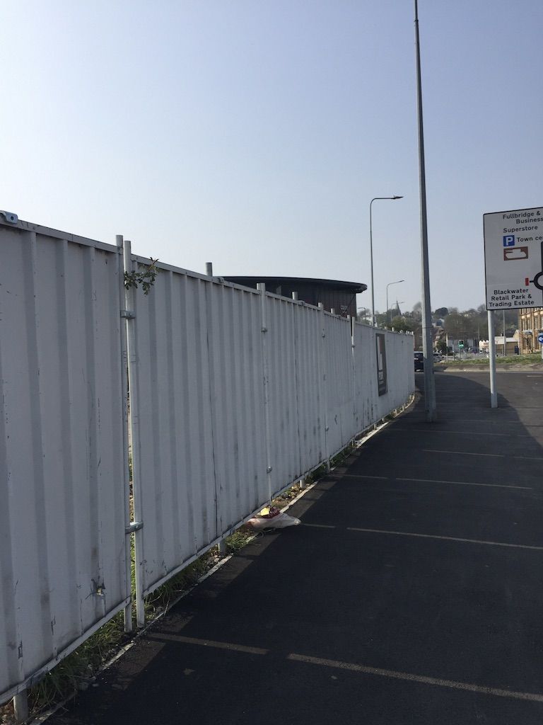 There is a white metal fence running along the left hand side of this image. It is made of corrugated metal, and has been ererected as a temporary measure to hide building works. It runs adjacent to the pavement.